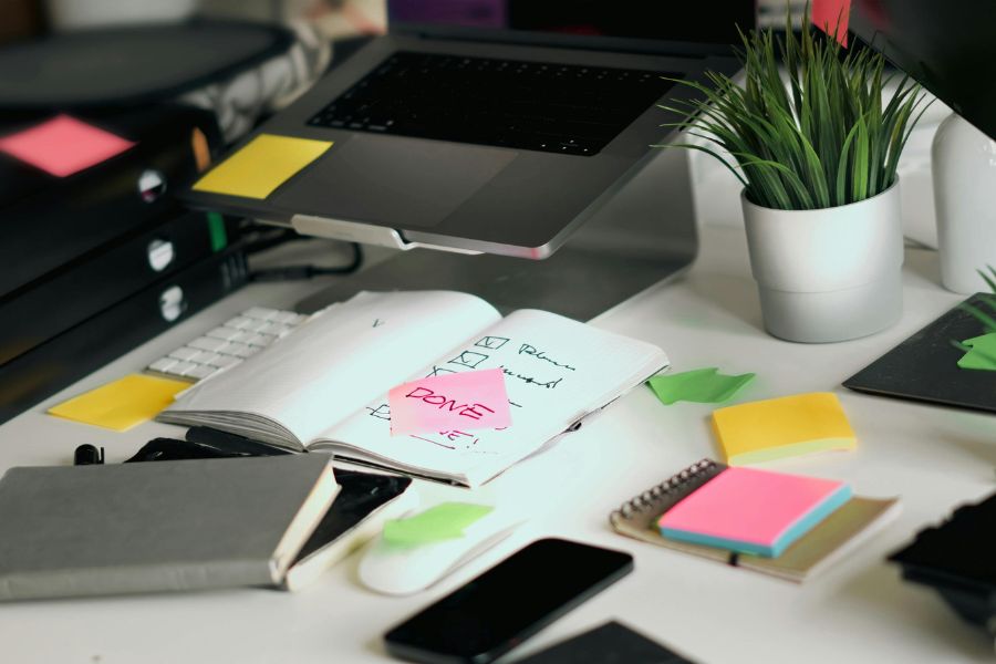 Un bureau encombré avec un ordinateur portable, des carnets, des blocs-notes adhésifs colorés et un post-it rose portant la mention "Done".